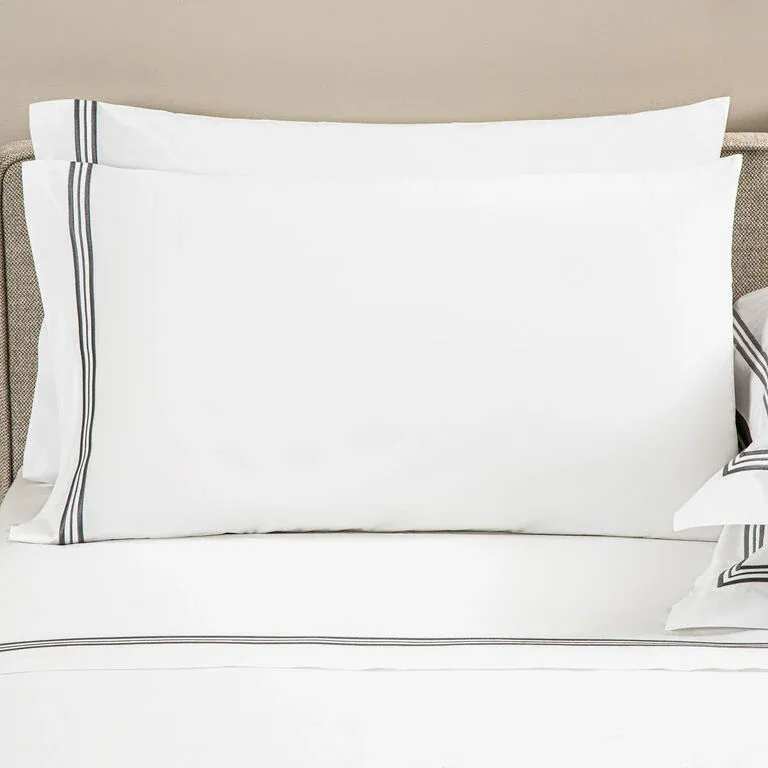A neatly made bed with Frette’s Triplo Bourdon pillowcase set in white cotton poplin, detailed with simple black and gray three-line embroidery along the borders.
