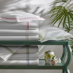A neatly arranged display of Amara Triple Pearl linens and pillows with ombre pearl-like strands of embroidery on a green shelf, accompanied by decorative vases and flowers.