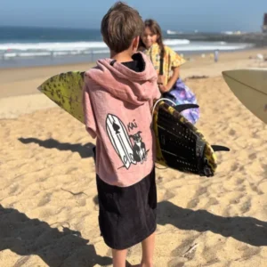 Enfant portant le poncho surf ALL-IN Kaï Surf Kid sur la plage, illustration Kaï et sa planche au dos