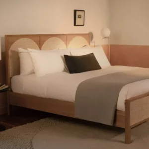 A bedroom with a contemporary wood and rattan bed made with LETTO Americano Cotton Percale sheets, a gray blanket folded at the foot of the bed, and a charcoal velvet throw pillow.